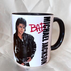 Puede incluir: Una taza de cerámica blanca con asa e interior negros, que presenta una impresión fotográfica de Michael Jackson con una chaqueta de cuero negra. El texto BAD está impreso en rojo sobre el nombre MICHAEL JACKSON, escrito verticalmente en negro.