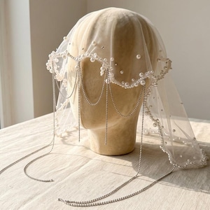 May include: A sheer ivory tulle veil draped over a mannequin head, featuring a scalloped edge adorned with white pearls of varying sizes. The veil is accented with delicate silver-toned metal chains that hang down in multiple draped strands.