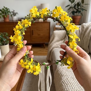 以下が含まれることがあります： 鮮やかな黄色の花と小さな緑の葉が、細く柔軟な茶色の蔓のようなベースに取り付けられた円形のフラワー冠。冠の下部からは、調整用に2本の長く細いオフホワイトのリボンが垂れ下がっています。