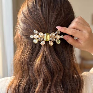 May include: A hair clip featuring a central rectangular yellow crystal surrounded by multiple teardrop-shaped crystals in shades of champagne, light yellow, and clear, all set on a metallic base and secured in long, wavy brown hair.