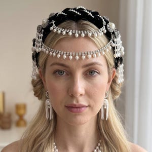 May include: A black velvet padded headband featuring a braided design adorned with white faux pearls and silver-toned crystal embellishments. The accessory includes dangling silver chains with pearl accents that drape across the forehead.