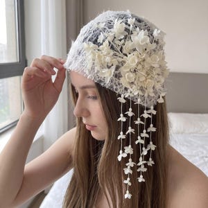 Puede incluir: Un tocado de encaje blanco que presenta un grupo de pequeñas flores blancas esculpidas en un lado. Varios hilos delicados de diminutos dijes florales blancos cuelgan del arreglo floral principal, creando un efecto de cascada sobre el cabello.