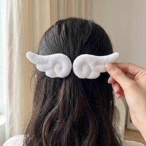 Puede incluir: Un par de pinzas para el cabello de felpa blanca con forma de alas de ángel estilizadas. Las alas de tela suave y texturizada presentan segmentos curvos y redondeados, diseñadas para llevarse en el cabello como un accesorio decorativo brillante.