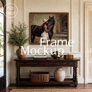 May include: A rectangular wall frame mockup featuring a painting of a person in a straw hat embracing a black horse. The frame is displayed above a dark wooden console table with drawers, a woven basket, a ceramic vase, and stacked books.
