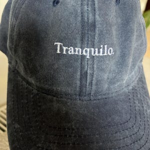 May include: A navy blue baseball cap with a washed, distressed texture. The front panel features the word Tranquilo embroidered in white, serif-style lettering. The cap includes a curved brim with visible stitching and eyelets on the crown.