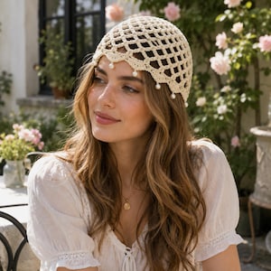 May include: A cream-colored crochet cap featuring an open diamond-shaped lattice pattern. The edge of the cap has a scalloped design with small, round, white faux pearls dangling from the points of the scallops, creating a delicate, textured look.