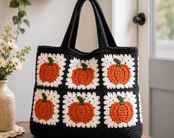 Puede incluir: Un bolso de ganchillo negro con un patrón de cuadrados blancos, cada uno con una calabaza naranja texturizada con un tallo verde. El bolso tiene dos asas negras resistentes y está construido con hilo, mostrando un diseño estacional.