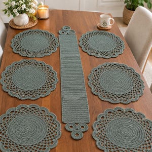 Op de afbeelding: Een set van zes saliegroene gehaakte ronde placemats en een bijpassende lange rechthoekige tafelloper met decoratieve cirkelvormige accenten aan beide uiteinden, voorzien van een dicht middenstuk en een geschulpte rand.