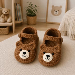May include: A pair of brown crochet slippers featuring a bear face design on the front, complete with small rounded ears, a cream-colored muzzle, black embroidered eyes, and a nose. Each slipper includes a strap secured with a small brown button.