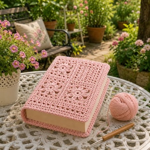 May include: A light pink crocheted book cover featuring a floral motif, accompanied by a matching ball of yarn and a wooden crochet hook. The cover is constructed with a detailed stitch pattern and fits snugly around the rectangular book.