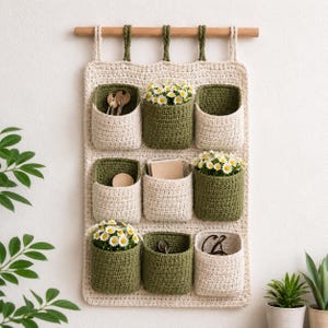 May include: A wall-hanging crochet organizer featuring nine pockets in alternating cream and olive green colors. The organizer is suspended from a wooden rod by five loops. The pockets contain small items including daisies, keys, and notebooks.