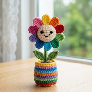 May include: A crocheted flower with a beige center, black bead eyes, and a smiling mouth. The petals are arranged in a rainbow pattern of pink, yellow, orange, red, green, blue, and purple. It has a green stem with two leaves in a striped pot.