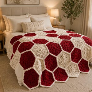 Op de afbeelding: Een handgemaakte gehaakte deken met een herhalend geometrisch patroon van in elkaar grijpende zeshoeken in afwisselende kleuren van donkerrood en crèmewit garen, gedrapeerd over een bed met een getextureerd oppervlak en geschulpte randen.