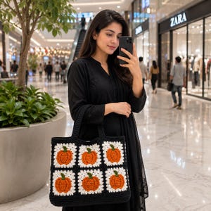 Puede incluir: Un bolso tote de ganchillo negro con una cuadrícula de seis motivos cuadrados. Cada cuadrado contiene una calabaza naranja texturizada con un tallo verde, enmarcada por hilo blanco. El bolso tiene dos asas negras resistentes y una forma rectangular, construida con un patrón de punto de hilo visible en toda la pieza.