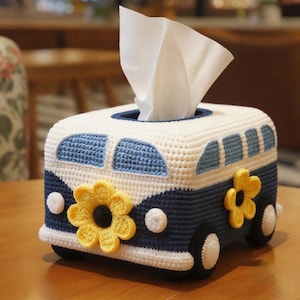 May include: A rectangular crochet tissue box cover shaped like a van, featuring a white top and dark blue bottom. It includes light blue windows, white circular headlights, black wheels with white centres, and yellow flower appliqués on the front and side.