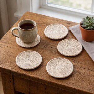May include: A set of five round, cream-colored crocheted coasters made from thick cord. One coaster supports a speckled ceramic mug filled with dark liquid, while the other four are arranged on a wooden surface alongside a small potted succulent.