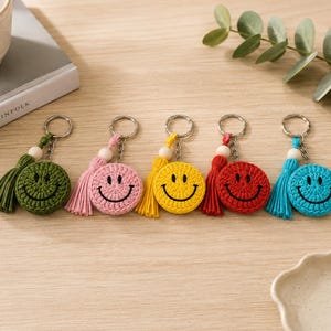 Könnte beinhalten: Fünf runde, gehäkelte Smiley-Schlüsselanhänger in Olivgrün, Hellrosa, leuchtendem Gelb, Rot und Türkis. Jeder Schlüsselanhänger verfügt über eine passende Garnquaste, eine kleine Holzperle und einen silberfarbenen Metallring zur Befestigung.