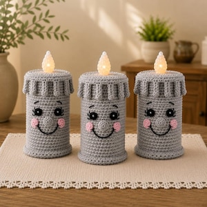 May include: Three cylindrical gray crochet candle covers featuring embroidered faces with black eyes, long eyelashes, pink cheeks, and a wide smile. Each cover is topped with a textured, off-white, flame-shaped LED light insert.