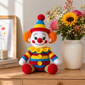 May include: A handmade crochet clown doll featuring a white face, red nose, and orange hair. It wears a blue, yellow, and red striped hat with a pom-pom, a yellow ruffled collar, and a matching striped body with two yellow buttons on the front.