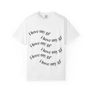 May include: A white short-sleeved t-shirt featuring the black text "i love my gf" printed six times in a repeating, staggered, and slightly curved pattern across the centre of the chest. The shirt has a crew neckline and a standard fit.