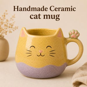 Peut inclure: Un mug en céramique jaune et violet en forme de chat avec des oreilles pointues, un nez rose et des moustaches. L'anse présente une petite empreinte de patte de chat rose. Le texte "Handmade Ceramic cat mug" est écrit en brun foncé au-dessus.