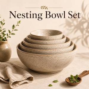 Pode incluir: Um conjunto de cinco tigelas empilháveis com acabamento bege salpicado, dispostas da menor para a maior. O texto "Nesting Bowl Set" está impresso acima. O conjunto inclui um vaso salpicado a condizer, um pano bege e uma colher de madeira.