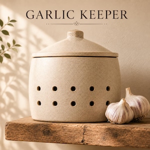 May include: A beige speckled ceramic garlic keeper with a lid and two rows of ventilation holes. The text GARLIC KEEPER is printed above the container. Two whole garlic bulbs are positioned to the right of the cylindrical storage vessel.