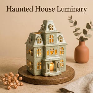 May include: A light green ceramic haunted house luminary with a speckled finish, featuring multiple arched windows and a cutout ghost shape. The structure is illuminated from within by a warm light, and the text Haunted House Luminary is displayed above.