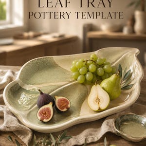 Puede incluir: Una bandeja de cerámica con forma de hoja y esmalte brillante en color crema y verde salvia. La bandeja muestra el texto LEAF TRAY POTTERY TEMPLATE y contiene uvas verdes, una pera entera, media pera y dos mitades de higo sobre una mesa.