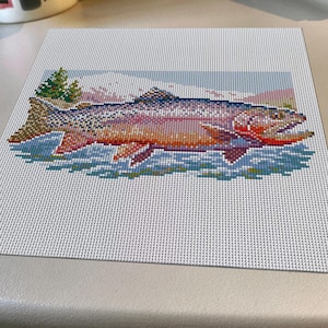 May include: A square needlepoint canvas featuring a colorful trout swimming in water. The fish displays shades of pink, orange, and blue, with a detailed tail and fins. In the background, there are green pine trees and a light blue sky, all rendered in a grid-like pattern on a white mesh background.