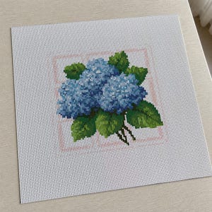 May include: A square piece of white needlepoint canvas featuring a cross-stitch design of a blue hydrangea flower cluster with green leaves. The floral pattern is centered within a thin, light pink square outline on the grid fabric.