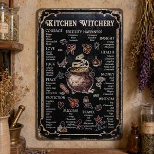 May include: A rectangular black metal sign titled Kitchen Witchery in white text. It features a central illustration of a steaming cauldron surrounded by lists of herbs and spices categorized by themes like Courage, Love, Luck, Peace, and Protection.