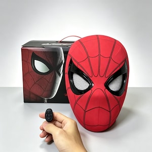 May include: A red Spider-Man mask featuring black web patterns and large white mesh eye lenses. The mask is positioned next to its original black box, which displays a graphic of the mask, and a small black remote control with five buttons.