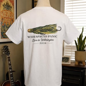 May include: A white short-sleeve t-shirt featuring a graphic of a green and yellow fishing lure on the back. Below the lure, the text reads WIDESPREAD PANIC, Live in Wilmington, and 2026. The shirt is displayed on a mannequin torso.