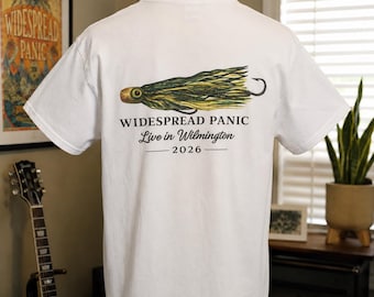 May include: A white short-sleeve t-shirt featuring a graphic of a green and yellow fishing lure on the back. Below the lure, the text reads WIDESPREAD PANIC, Live in Wilmington, and 2026. The shirt is displayed on a mannequin torso.