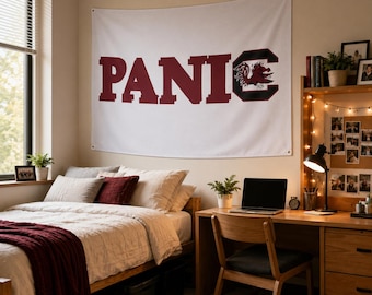 May include: A white rectangular wall tapestry featuring the maroon text PANIC with a stylized maroon gamecock mascot graphic integrated into the letter C. The fabric banner is mounted on a wall above a bed and desk in a bedroom setting.