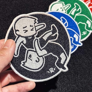 Könnte beinhalten: Runde, gestickte Aufnäher mit einer Cartoon-Illustration eines Ringkampfes. Die Aufnäher haben einen schwarzen Hintergrund und einen weißen Rand. Zusätzliche Aufnäher in Blau, Grün und Rot sind sichtbar. Geeignet für Kleidung.