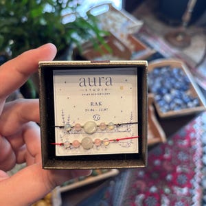 Puede incluir: Dos pulseras de cuentas presentadas en una tarjeta de marca dentro de una caja. La pulsera superior tiene un cordón negro y la inferior uno rojo. Ambas cuentan con una piedra central verde pálido flanqueada por cuentas rosas e iridiscentes.
