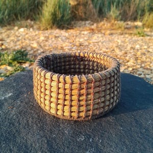 May include: A small, round, open-top basket constructed from light tan dried grass or straw coils. The coils are bound together by thin, dark brown thread or cord, creating a visible vertical grid pattern around the exterior of the vessel.