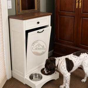 May include: A white wooden pet feeding station with a dark wood top, a single drawer with a black knob, and a tilt-out cabinet door featuring a black handle. The unit includes two integrated stainless steel bowls, one containing dry food.