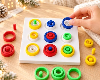 Può includere: Una tavola da gioco quadrata bianca con una griglia tre per tre di rientranze circolari. La tavola presenta vari anelli e pioli a incastro nei colori primari rosso, blu, giallo e verde, disposti nella griglia e sparsi attorno.
