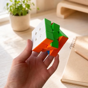 Puede incluir: Un cubo de rompecabezas 3x3 con una cuadrícula de paneles cuadrados en colores blanco, verde y naranja. El cubo se sostiene en una posición que muestra tres de sus lados, revelando un estado parcialmente resuelto con segmentos de colores.