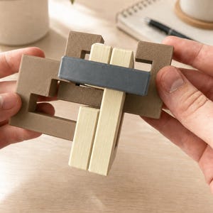 Könnte beinhalten: Ein 3D-gedrucktes Puzzle aus ineinandergreifenden geometrischen Teilen. Die Struktur besteht aus einem großen, braunen, U-förmigen Rahmen, zwei vertikalen cremefarbenen rechteckigen Stäben und einem horizontalen dunkelgrauen Stab.
