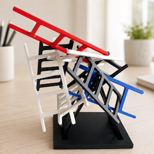 Op de afbeelding: Een sculpturale opstelling van meerdere miniatuurladders in rood, wit, zwart en blauw, gestapeld en in verschillende hoeken in evenwicht gebracht. De structuur wordt ondersteund door een vierkante zwarte basis.