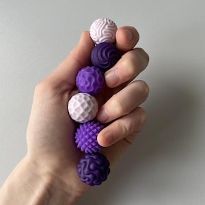 May include: A set of six small, spherical sensory balls in varying shades of purple and lavender. Each ball features a distinct tactile surface pattern, including swirls, geometric grids, and raised spikes, designed for sensory stimulation.