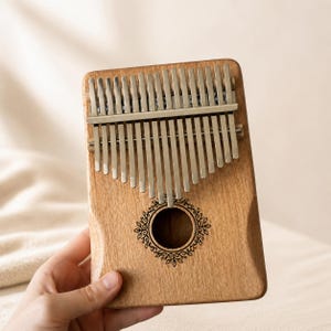 May include: A wooden kalimba musical instrument featuring seventeen silver-colored metal tines arranged in a row. The rectangular wooden body has a central circular sound hole decorated with a black engraved floral vine pattern around the edge.