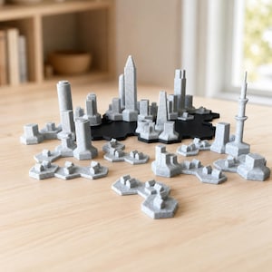 May include: A set of gray, speckled, 3D-printed city skyline building models arranged on a black, hexagonal base. The collection includes various architectural structures, including skyscrapers, towers, and smaller building blocks with geometric shapes.