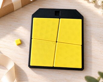 Può includere: Una cornice rettangolare nera con un foro di sospensione superiore contenente quattro piastrelle quadrate giallo brillante con una sottile trama geometrica. Un singolo piccolo pezzo quadrato giallo è posizionato a sinistra della cornice.