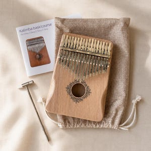 May include: A light brown wooden kalimba with seventeen metal tines and a circular sound hole featuring a decorative floral engraving. The set includes a small metal tuning hammer, a beige drawstring storage pouch, and a white instruction booklet.