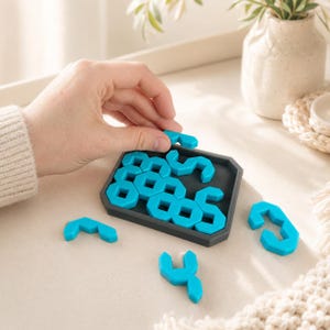 May include: A brain teaser puzzle consisting of a dark gray rectangular tray and multiple bright turquoise geometric pieces. Several pieces are fitted into the tray, while three additional turquoise pieces are placed on the surface nearby.